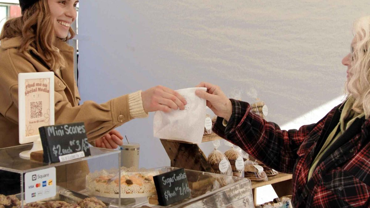 A market vendor handing a paper bag of baking to a customer