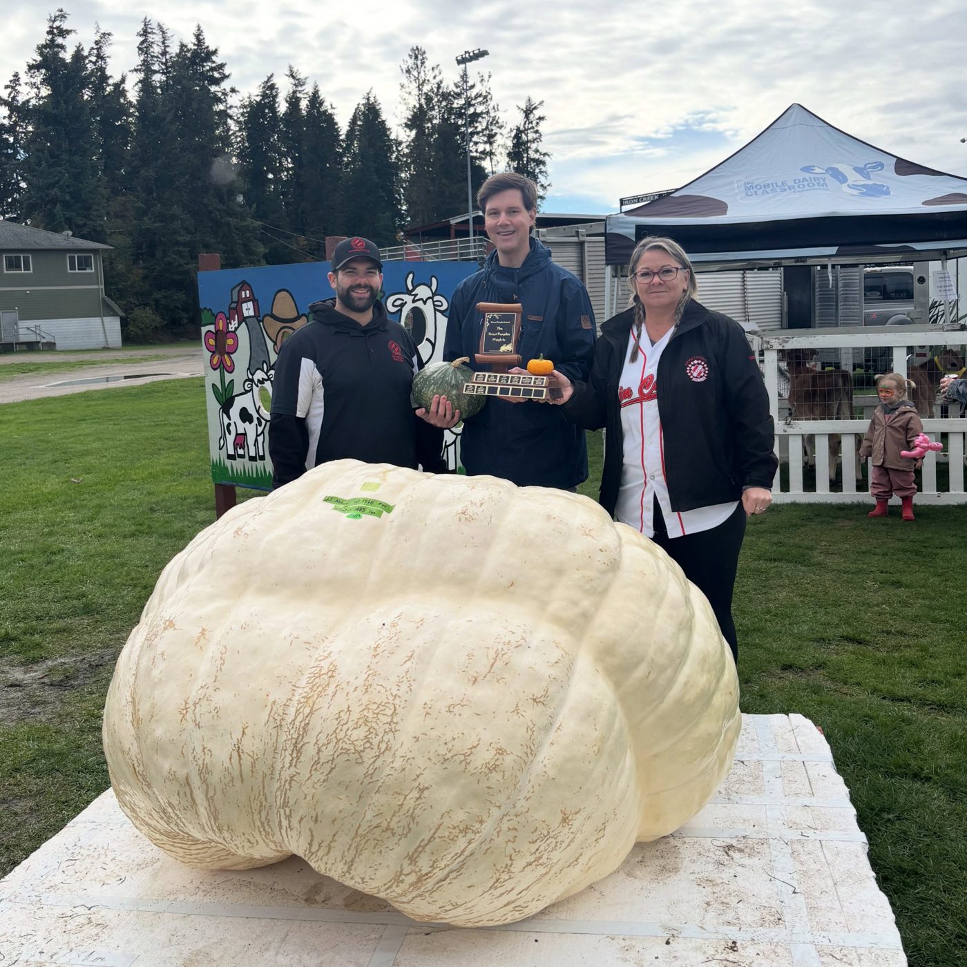 Jordan Abbate's 1,485 lb giant pumpkin at the 2026 Great Pumpkin Weigh-In, with the trophy and two Chamber volunteers