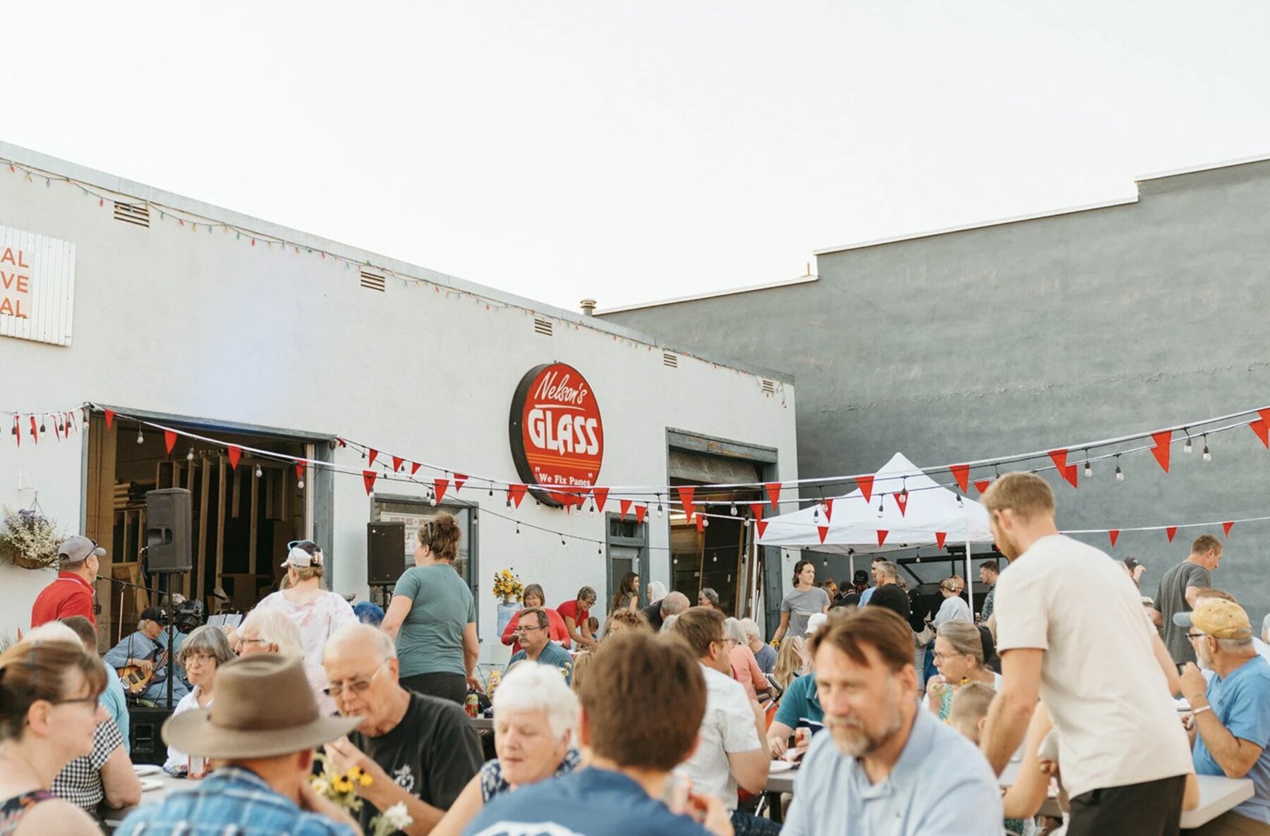 People gather at outdoor tables outside Nelson’s Glass, beneath red pennants and string lights.